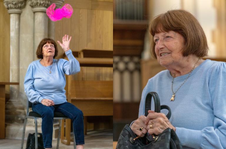 An elderly woman in a light blue jumper sits on a chair in a church, reaching up toward a pink translucent fabric floating above her raised hand. A close-up shows her smiling face with short brown hair, wearing a necklace, and holding a black handbag.