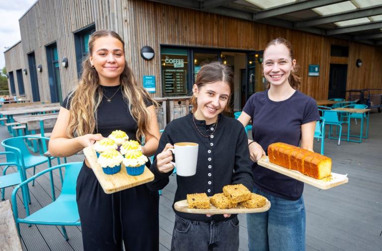 DP Hospitality Team at The Coffee Deck cafe at Dinton Pastures. They are holding a selection of cakes and a latte.