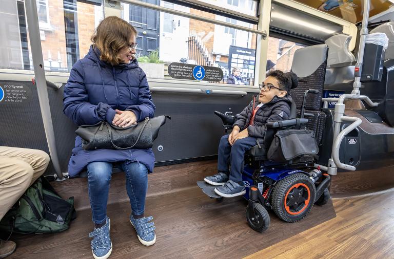 A woman in a blue puffer jacket sits on a bus seat with a black bag on her lap. A man in a motorised wheelchair sits nearby.