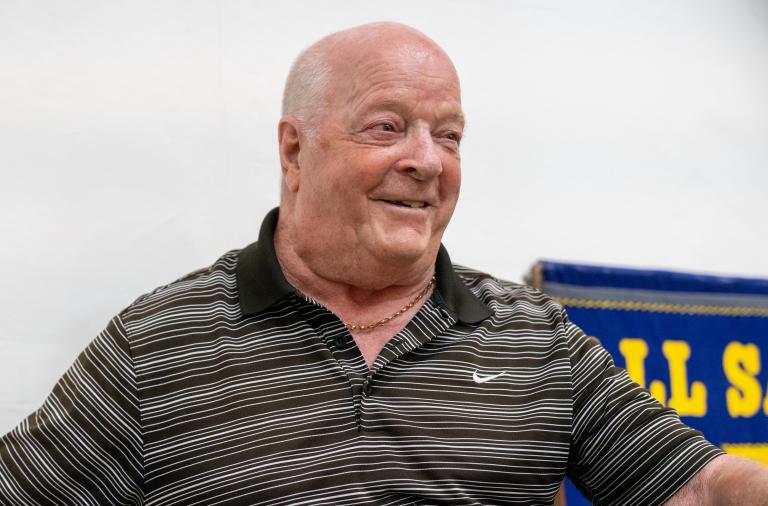 A smiling, older man in a dark striped polo shirt with a small white Nike swoosh and a thin gold necklace. The light wall behind him shows a blue object to the right.