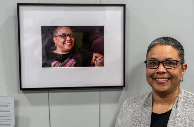 A smiling person with short grey hair and glasses stands beside their framed portrait on a light-grey wall. The portrait shows them smiling against a dark background.