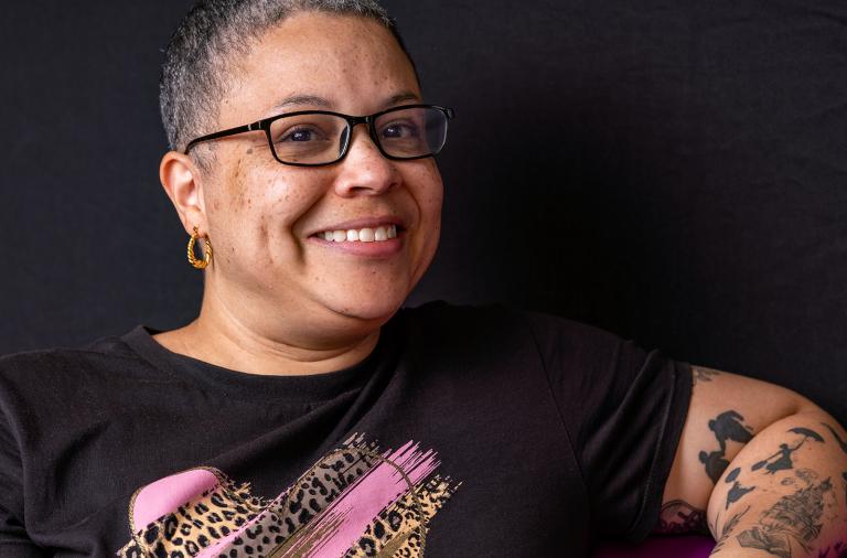 Smiling person with short-cropped hair and glasses, wearing a black T-shirt with a pink leopard-print graphic. They have a gold hoop earring and visible tattoos on the left arm, against a dark backdrop.