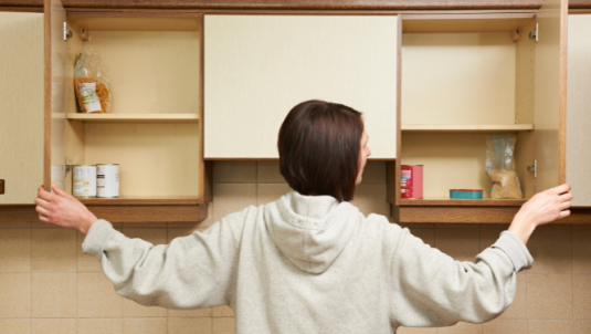 A woman opens two kitchen cupboards showing a few tinned goods left at the back of the cupbard