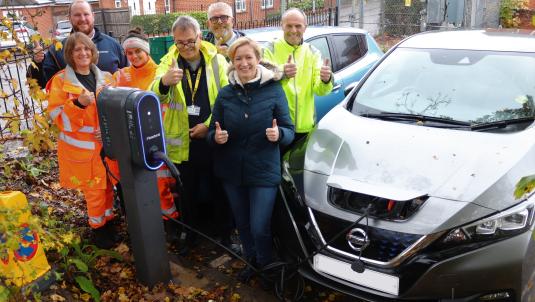 A group of men and women, many in high vis gear, cheer and give the thumbs up next to two cars charging side by side in a car park