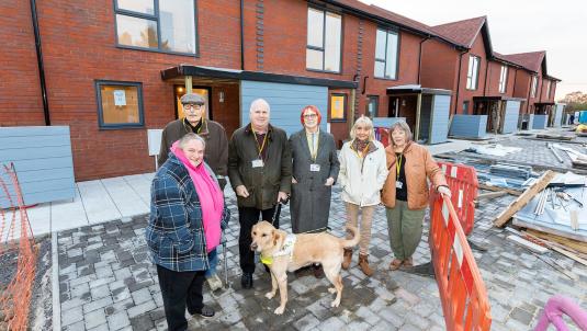 Six tenant volunteers and a guide dog stand outside a new row of council houses nearing construction