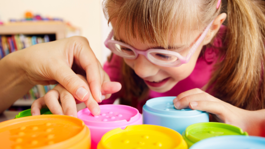 A girl leans over a set of textured stacking cups, with an adult helping her to feel the surface