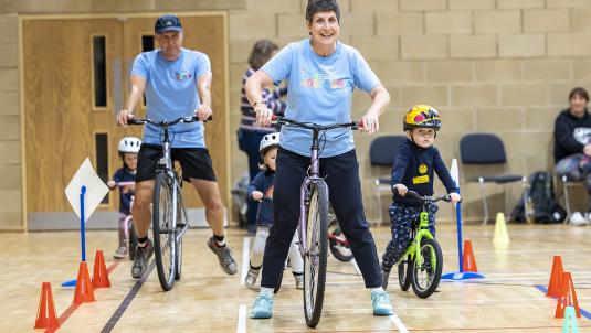 Photo of Jill Bissell riding a bike around an obstacle course in a sports hall as little children watch and learn