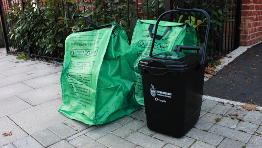 Photograph of two full green recycling bags and a black food waste bin at the kerbside in front of a house