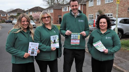 Three women and a man wearing dark green council jackets, standing in a residential street and holding up information leaflets