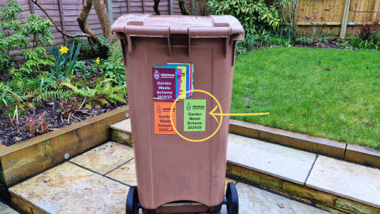 A brown garden waste bin with stickers in a garden