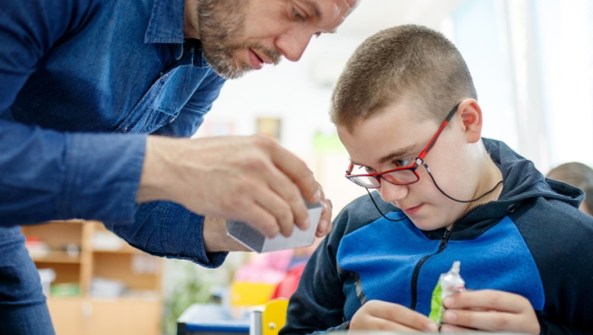 A boy is in a classroom working on a piece of craft with the help of an adult male