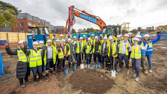 Workers in hi-vis and hard hats celebrate and raise hands on muddy site with an orange excavator bearing a Union Jack behind them.