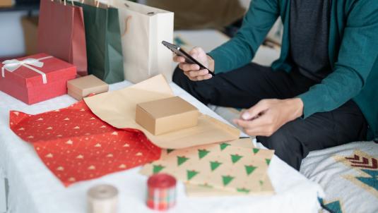Person using phone near gifts and wrapping paper on a bed.