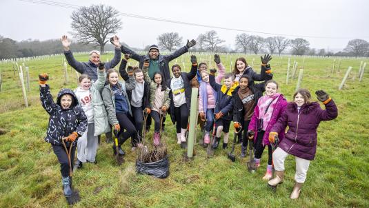 Schoolchildren planting saplings in a field at the Covid Memorial Woodland, holding spades and celebrating their work.