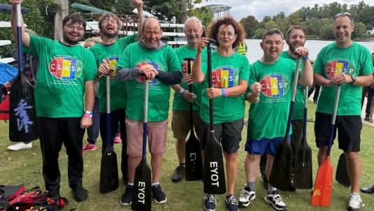 Eight people in matching green CLASP t-shirts stand on grass by a lake, each holding a canoe paddle. They smile and pose for the camera with boats and trees in the background.