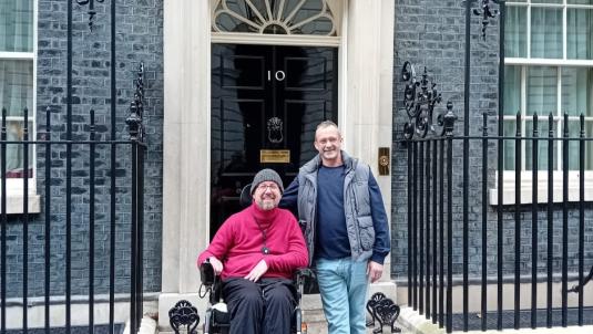 Two men stand outside number 10 Downing Street. One man sits in a wheelchair, while the other stands beside him, with wrought-iron railings along the doorway.