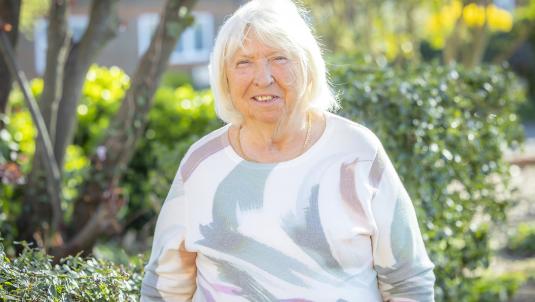 A woman with short white hair stands in a sunny garden, wearing a pastel abstract-patterned sweater. Green shrubs and blurred yellow flowers are in the background.