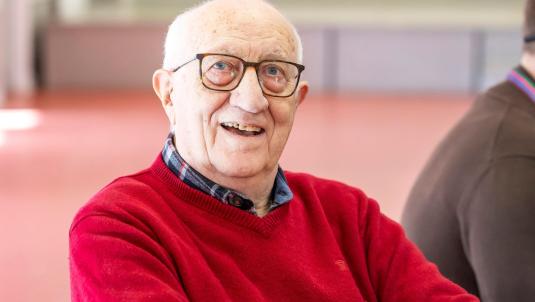 An elderly man with white hair and glasses smiles at the camera. He wears a red sweater over a plaid shirt, and the background is a softly blurred indoor space.