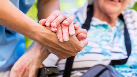 Two hands are clasped in a supportive gesture. In the blurred background, an elderly person wearing a blue floral shirt sits in a wheelchair and smiles.