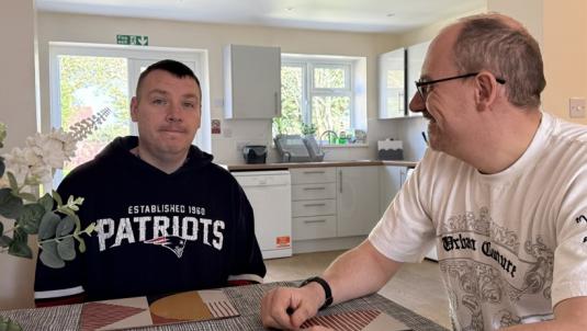 Two men sit across from each other at a dining table in a bright, modern kitchen. The man on the left wears a black Patriots hoodie; the man on the right wears a white graphic T‑shirt and glasses, and they smile at each other.