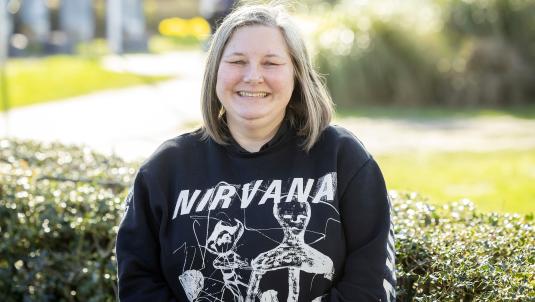 A person with shoulder-length blond hair smiles outdoors. They wear a black Nirvana sweatshirt with a skeletal graphic, standing in front of hedges on a sunny day.