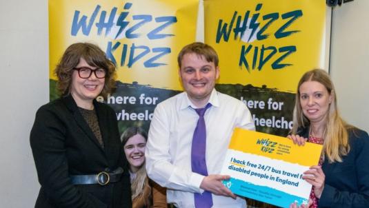 Three people pose in front of yellow Whizz-Kidz banners. The centre person in a white shirt and purple tie is flanked by a woman with glasses on the left and a woman on the right holding a sign supporting free 24/7 bus travel for disabled people in England.