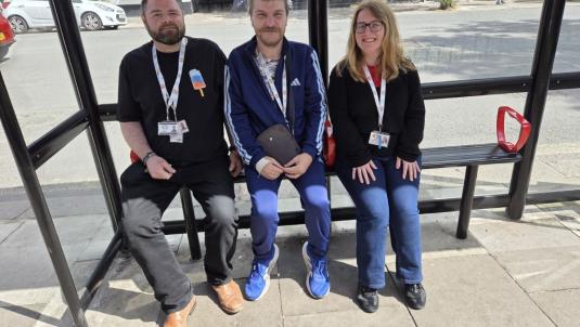 Three people sit on a bench inside a glass bus stop shelter on a town street. Left to right: a bearded man in a black T-shirt and tan boots, a man in a blue tracksuit holding a dark folder, and a smiling woman with glasses in a black jumper and jeans.