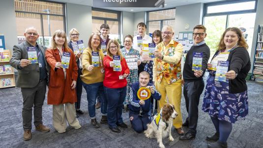 A group of adults pose in a library reception area, all holding blue 'Safe Places' leaflets. A dog in a vest sits in front; a 'Reception' sign and windows are in the background.