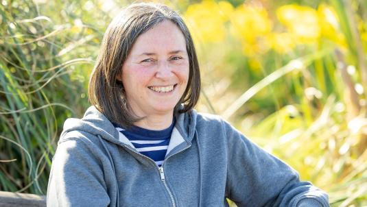 Smiling woman with short dark bob, wearing a blue striped shirt under a grey zip hoodie. Outdoors, sunlit greenery provides a blurred yellow-green background.