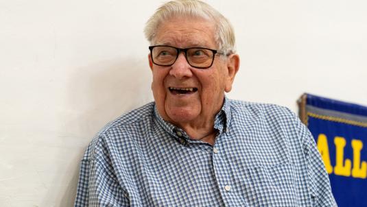 An elderly man with white hair and black-framed glasses smiles. He wears a blue-and-white checkered shirt; a blue banner with yellow letters is visible in the background on the side.