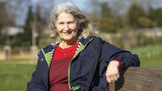 An older woman with grey hair sits on a wooden park bench, smiling at the camera. She wears a navy jacket over a red jumper, with a bright green zipper and a crossbody bag, in a sunny park with blurred trees in the background.
