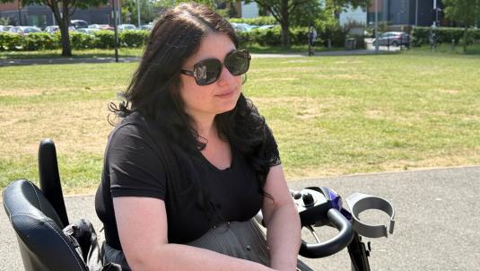 A woman with long dark hair wearing large sunglasses and a black T-shirt sits on a mobility scooter in a sunny park. In the background, there is grass, trees, and buildings with parked cars along a street.