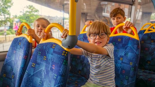 Two young boys and one young girl sitting on the back seats of a bus while smiling at camera