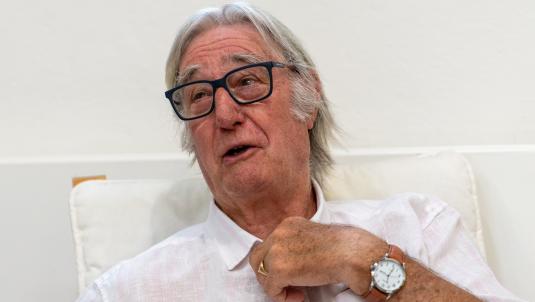An older man with grey hair and dark rectangular glasses sits on a light chair against a pale wall. He wears a light shirt and a brown leather wristwatch on his left wrist, and he appears to be speaking.