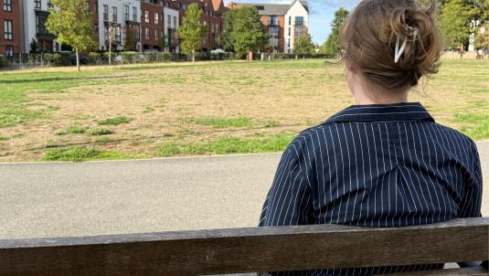 Back view of a person with light brown hair tied with a hair clip, wearing a navy pinstriped shirt, seated on a wooden bench in an urban park.