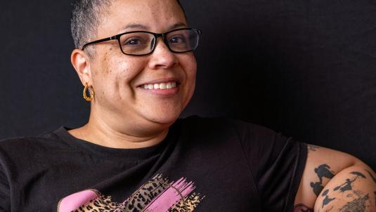 Smiling person with short-cropped hair and glasses, wearing a black T-shirt with a pink leopard-print graphic. They have a gold hoop earring and visible tattoos on the left arm, against a dark backdrop.