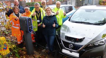 A group of men and women, many in high vis gear, cheer and give the thumbs up next to two cars charging side by side in a car park