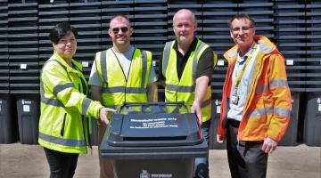 Four people in high vis jackets gather around a new bin and smile to the camera, with several huge stacks of new bins behind them
