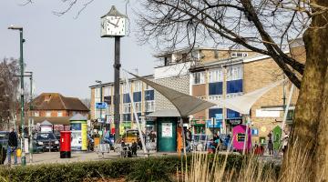 Woodley precinct on a busy sunny day with its clock tower and canopy in background, with trees and long grass in foreground
