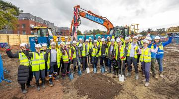 Workers in hi-vis and hard hats celebrate and raise hands on muddy site with an orange excavator bearing a Union Jack behind them.