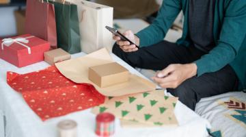 Person using phone near gifts and wrapping paper on a bed.