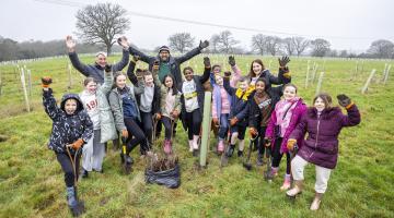 Schoolchildren planting saplings in a field at the Covid Memorial Woodland, holding spades and celebrating their work.