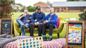 Three men sit on a lime-green sofa on grass, laughing. A chalkboard sign and a Public Living Room poster to each side.
