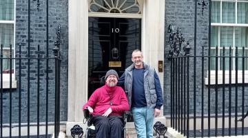 Two men stand outside number 10 Downing Street. One man sits in a wheelchair, while the other stands beside him, with wrought-iron railings along the doorway.