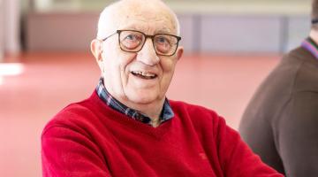 An elderly man with white hair and glasses smiles at the camera. He wears a red sweater over a plaid shirt, and the background is a softly blurred indoor space.