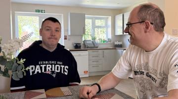 Two men sit across from each other at a dining table in a bright, modern kitchen. The man on the left wears a black Patriots hoodie; the man on the right wears a white graphic T‑shirt and glasses, and they smile at each other.