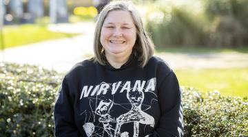 A person with shoulder-length blond hair smiles outdoors. They wear a black Nirvana sweatshirt with a skeletal graphic, standing in front of hedges on a sunny day.