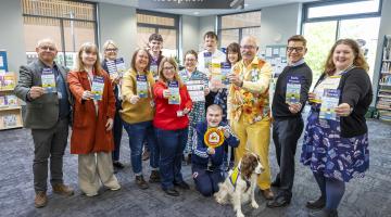 A group of adults pose in a library reception area, all holding blue 'Safe Places' leaflets. A dog in a vest sits in front; a 'Reception' sign and windows are in the background.