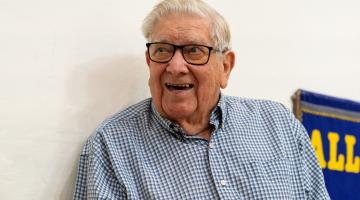 An elderly man with white hair and black-framed glasses smiles. He wears a blue-and-white checkered shirt; a blue banner with yellow letters is visible in the background on the side.