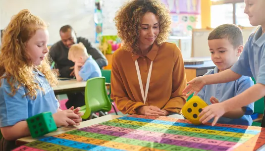 A teacher playing a maths game with 3 pupils, using a number grid and large dice